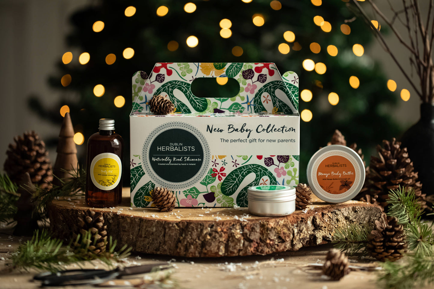 Herbalists New Baby Collection gift set on a wooden surface with decorative lights and pinecones in the background.
