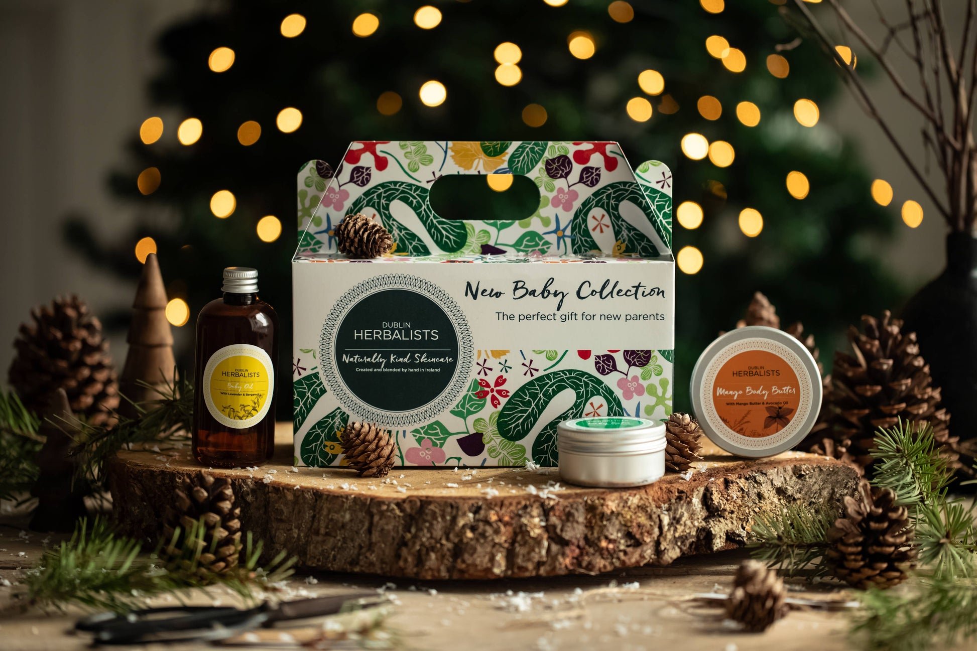 Herbalists New Baby Collection gift set on a wooden surface with decorative lights and pinecones in the background.